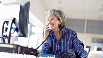 Eine lächelnde Frau sitzt am Schreibtisch vorm Computer und telefoniert.