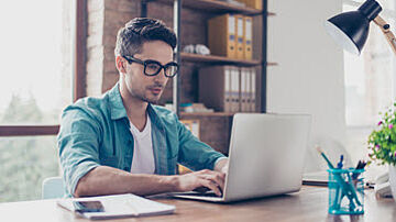 Junger Mann mit Brille sitzt am Schreibtisch und arbeitet am Laptop.