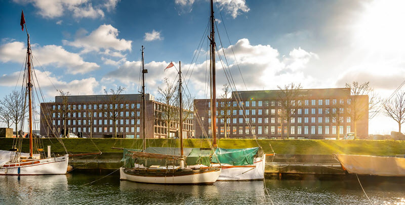 Drei historische Segelboote an einem Hafenkai. Dahinter zwei moderne Bürogebäude aus Backstein sowie blauer Himmel und Wolken.