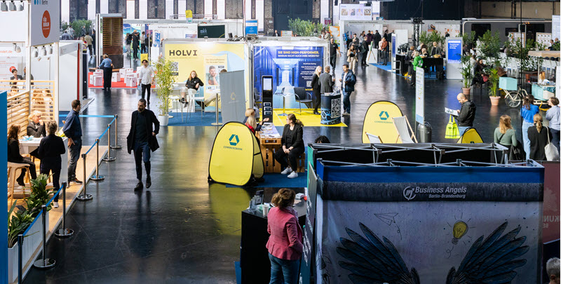 Personen gehen durch Messehalle mit Messeständen und Plakataufstellern. - Link auf Seite: Gründungsfrühstück deGUT