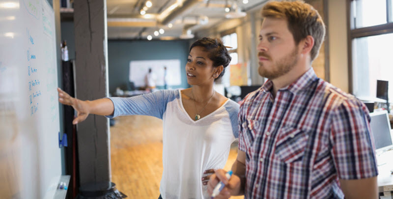 Gründen. Jetzt. – Start Up! Ein Mann und eine Frau stehen in einem Großraumbüro vor einem Flipchart. Die Frau zeigt mit der Hand auf das Flipchart. - Link auf Seite: Gründen. Jetzt. – Start Up!
