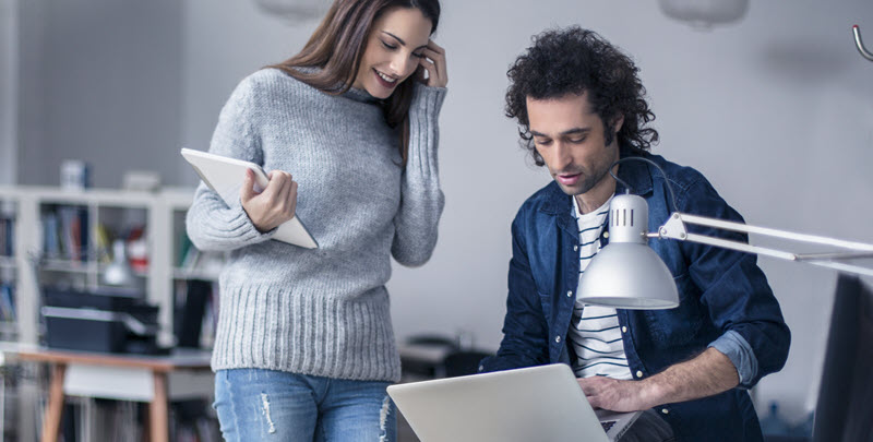 Eine Frau und ein Mann in einem Büro. Sie steht und hält ein Tablet in der rechten Hand. Er sitzt auf dem Schreibtisch mit einem Laptop auf seinem Bein.