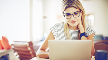 Junge blonde Schülerin mit Brille sitzt am Schreibtisch und arbeitet am Laptop