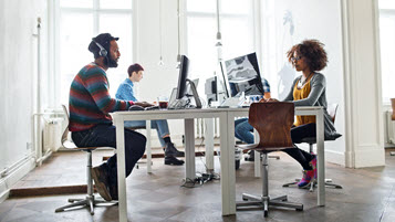 Vier Kollegen sitzen gemeinsam im Büro und arbeiten am Computer, einer von ihnen trägt Kopfhörer