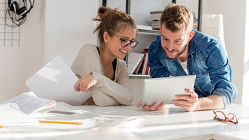 Junge Frau und junger Mann am Schreibtisch schauen zusammen auf ein Tablet.