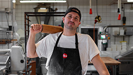 Mann mit Schürze und Kappe steht in einer Bäckerei oder Backstube und trägt ein Brot auf der Schulter. Im Hintergrund sind Arbeitsflächen und Backgeräte zu sehen.