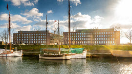 Drei historische Segelboote an einem Hafenkai. Dahinter zwei moderne Bürogebäude aus Backstein sowie blauer Himmel und Wolken.