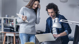 Eine Frau und ein Mann in einem Büro. Sie steht und hält ein Tablet in der rechten Hand. Er sitzt auf dem Schreibtisch mit einem Laptop auf seinem Bein.