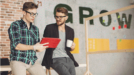 Zwei junge Männer lehnen an einem Tisch in einem Büro, einer von ihnen hält ein Tablet in Händen.