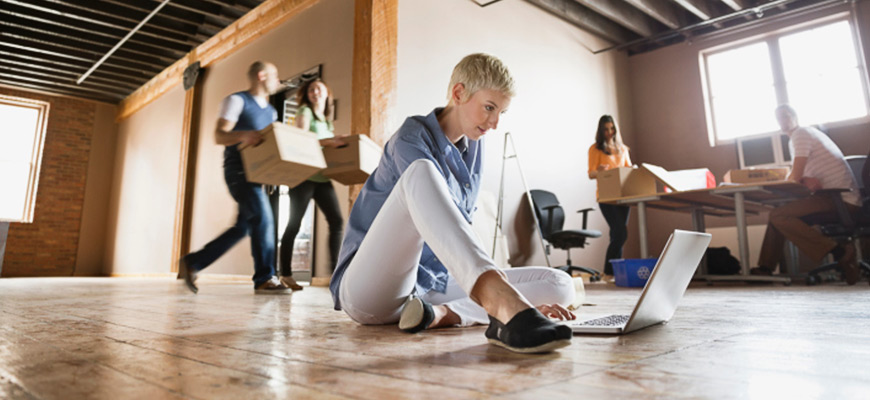 Junge Menschen richten ein neues Büro ein und eine Frau sitzt mit ihrem Laptop auf dem Boden