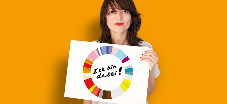 Eine junge Frau mit dunkelbraunen Haaren schaut freundlich in die Kamera und hält ein Plakat in der Hand, auf dem "Ich bin dabei!" steht.